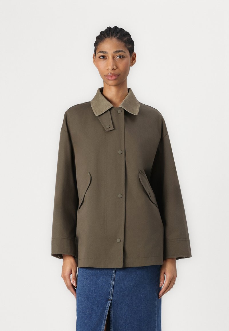 Olivově zelená bunda s měkkým béžovým límcem, patentkami, bočními kapsami a volným střihem. Materiál má hladkou, matnou texturu.