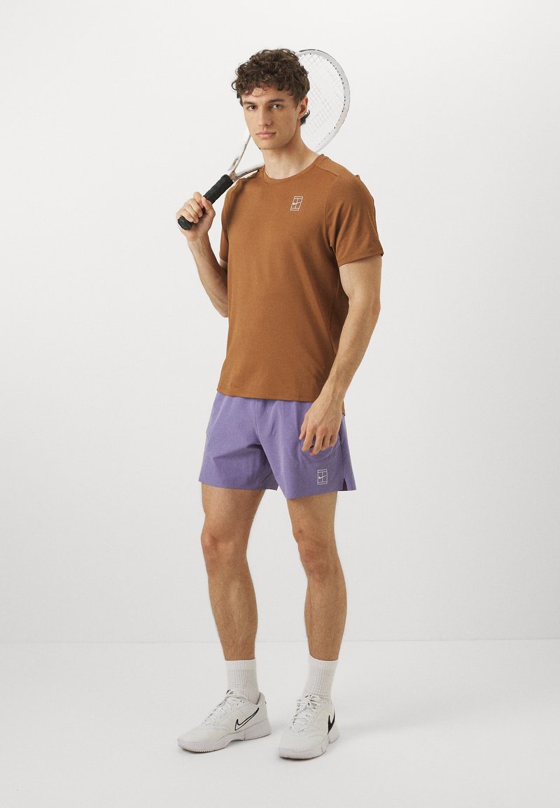 Braunes kurzärmliges Hemd, lila Shorts, weiße Turnschuhe. Hat ein Logo auf dem Hemd und den Shorts. Hält einen Tennisschläger in einer neutralen Pose.