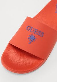 Chancla de goma roja con una amplia correa que lleva el logotipo "GUESS" en relieve y un gráfico de una palmera morada. Plantilla texturizada para un buen agarre.