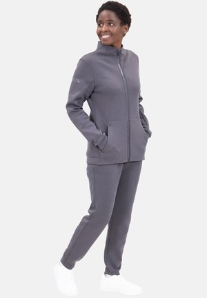 Graue Zip-Jacke mit Seitentaschen und passender Hose. Aus weichem Stoff gefertigt, hat einen hohen Kragen und Rippbündchen. Getragen mit weißen Turnschuhen.