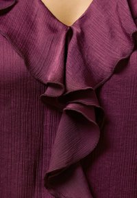 Burgunderfarbene Bluse aus strukturiertem Stoff mit vertikalen Falten und einem rüschierten V-Ausschnitt, der sanfte Drapierungen und glatte Akzente betont.