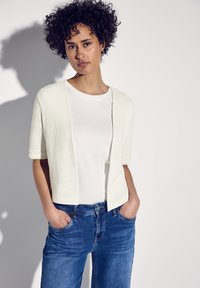 Kortärmad, öppen cardigan i strukturerad vit tyg, bärs över en enkel vit t-shirt och blå jeans, händerna i fickorna.
