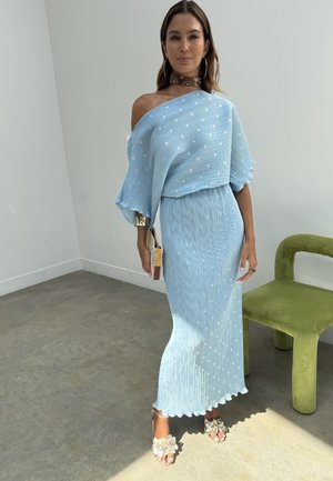 Žena stojící uvnitř, oblečená v světle modrých šatech s odhalenými rameny a bílými puntíky, držící pletené psaníčko, vedle zeleného sametového křesla.