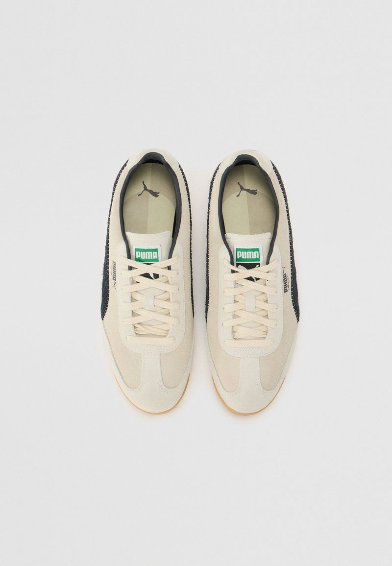 Sneaker bianche e nere con finitura in suede. Presentano un lato testurizzato, lacci beige, logo Puma verde sulla linguetta e suola in gomma.