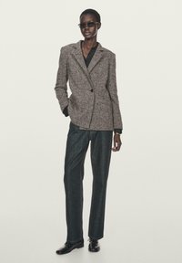 Blazer grigio a spina di pesce con un solo bottone, abbinato a pantaloni in denim scuri e scarpe nere. Il modello indossa occhiali da sole slim.