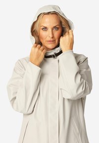 Donna con capelli biondi che indossa un impermeabile grigio chiaro, che aggiusta il cappuccio con entrambe le mani, espressione neutra, sfondo semplice e chiaro.