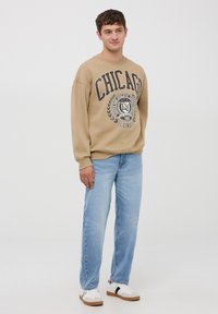 Felpa beige con grafica "CHICAGO", design casual. Jeans dritti azzurri chiari e scarpe da ginnastica bianche con accenti neri. Sfondo semplice.