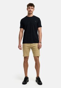 Sort bomulds t-shirt med brystlomme, parret med khaki shorts. Modellen bærer sorte sneakers og står foran en ensfarvet hvid baggrund.