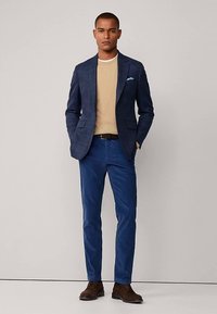 Marineblauwe geruite blazer over een beige trui, gecombineerd met blauwe chino's en bruine schoenen. De blazer heeft twee zakken en een pochet.
