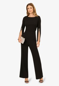 Černý overal s dlouhými rukávy a otevřenými stranami, ozdobený perlovými akcenty. Rozšířené kalhoty a přiléhavý pas vytvářejí elegantní siluetu.