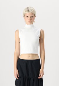 Hvit ermeløs cropped topp med høyt krage og glatt tekstur. Har en søm over brystet og en subtil logo nederst.