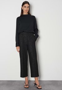 Zwarte lange mouwen top gecombineerd met zwarte wijde broek met een sprankelende textuur. Model draagt zwarte sandalen met hakken en eenvoudige oorbellen.