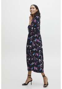 Robe noire jusqu'aux chevilles avec des motifs floraux en rose, bleu et blanc. Elle possède des manches longues et un design en couches fluide. Portée avec des talons noirs.