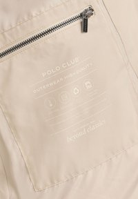 Beżowa kieszeń kurtki z czarnym zamkiem błyskawicznym, oznaczeniami Polo Club oraz ikonami wskazującymi na dodatkowe ocieplenie, wodoodporność, poliester z recyklingu, trwałość i możliwość prania.