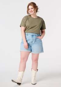 Olijfgroen T-shirt met korte mouwen en bloemenborduursel op de schouders, gecombineerd met lichtblauwe denim shorts en witte cowboylaarzen.