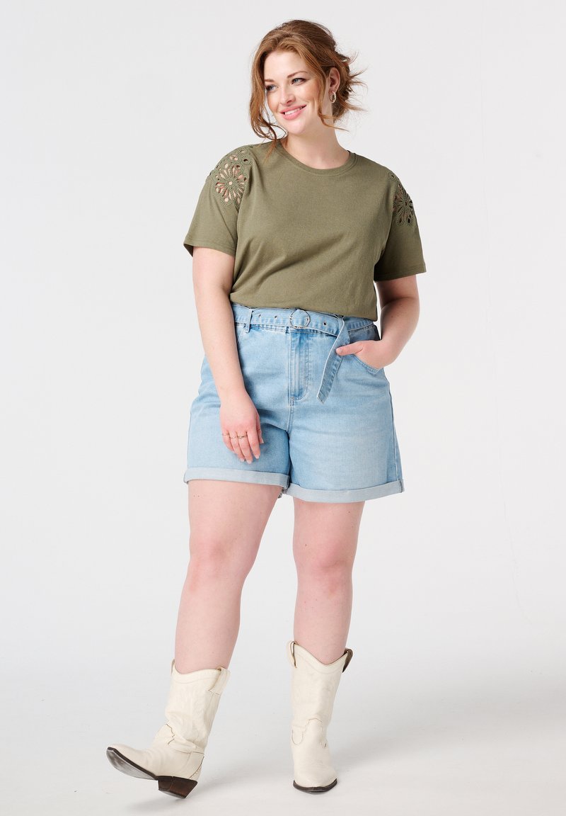 Olijfgroen T-shirt met korte mouwen en bloemenborduursel op de schouders, gecombineerd met lichtblauwe denim shorts en witte cowboylaarzen.