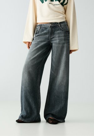 Personne portant un jean noir délavé à jambes larges associé à des chaussures en mesh noires et à un haut à manches longues crème avec texte vert.