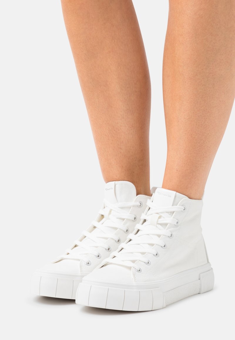Witte hoge sneakers met dikke rubberen zolen, gedragen op blote benen tegen een eenvoudige lichte achtergrond.