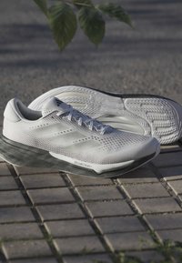 Valkoiset ja harmaat Adidas-juoksukengät, joissa on teksturoidut pohjat, asetettuna päällystetylle maalle vihreiden lehtien alle.