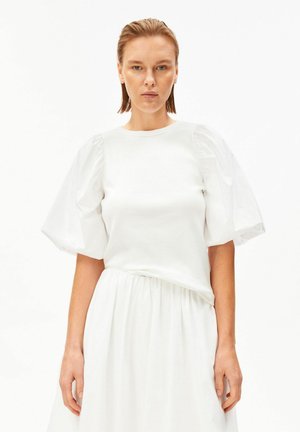 Femme aux cheveux plaqués en arrière portant une robe blanche avec des manches courtes bouffantes, debout devant un fond blanc.