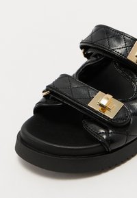 Sandalo nero con tomaia in patinato trapuntato, due cinturini regolabili, dettagli in metallo dorato e suola spessa e testurizzata.