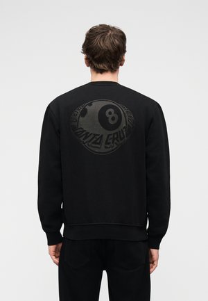 Férfi fekete pulóverben, amelynek hátulján nagy, sötétszürke 8-as golyó és "Santa Cruz" felirat látható, egyszínű háttér előtt áll.