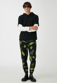 Svart kortärmad hoodie över långärmad vit tröja, parat med svarta byxor med gröna grafiska tryck och svarta stövlar.