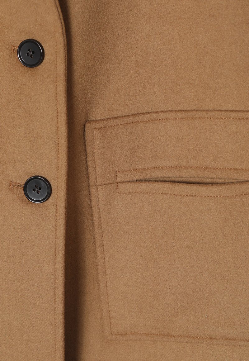 Cappotto in misto lana di colore beige, esposto con due bottoni neri, una tasca sul petto, una texture liscia e dettagli di cuciture pulite.