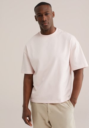 Mann mit kurzen Haaren, der ein hellrosa übergroßes T-Shirt und beige Hosen trägt, steht mit einer Hand in der Tasche vor einem schlichten Hintergrund.