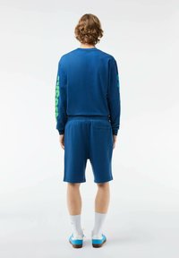 Camisa azul de manga larga con detalle del logo en verde, combinada con pantalones cortos azules. Los zapatos son azules con acentos blancos y se llevan calcetines blancos.