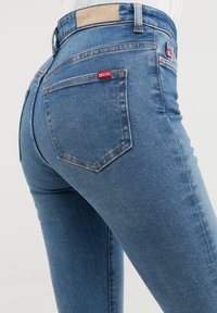 Mėlyni džinsai su prigludusia forma, turintys aukštą liemenį, dvi užpakalines kišenes ir mažą raudoną prekės ženklo etiketę dešinėje kišenėje.