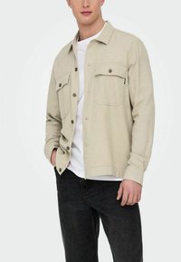 Veste en tissu beige avec col, deux poches avant et fermeture à boutons, portée par-dessus un t-shirt blanc et un jean foncé.