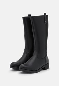 Par svart höga läderstövlar med låga klumpiga klackar och dragflikar, visade mot en enkel ljus bakgrund.