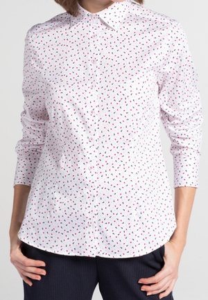 Weißes Hemd mit kleinen roten und schwarzen Herzmustern, lange Ärmel, ungeknöpft über dunkler Hose getragen.