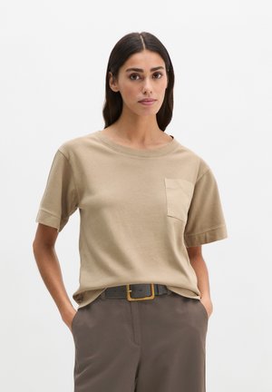 Kobieta o długich ciemnych włosach, ubrana w beżową koszulę z krótkim rękawem i kieszenią na piersi, wpuszczoną w ciemne spodnie z czarnym paskiem i złotą klamrą.