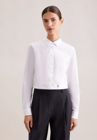 Kobieta o ciemnych, spiętych do tyłu włosach, ubrana w dopasowaną białą koszulę zapinaną na guziki i wysokowalne czarne spodnie, na jednolitym tle.