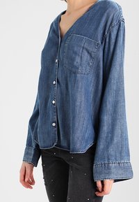 Femme portant une chemise ample en jean bleu boutonnée avec une poche poitrine et de larges poignets, associée à un pantalon noir orné de clous.