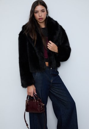 Sort cropped jakke i kunstpels med en bred krave, kombineret med mørkeblå high-waisted denimjeans og en lille mørkerød håndtaske.