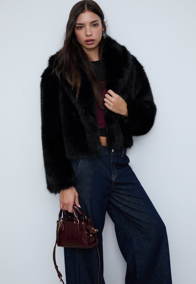 Sort cropped jakke i kunstpels med en bred krave, kombineret med mørkeblå high-waisted denimjeans og en lille mørkerød håndtaske.