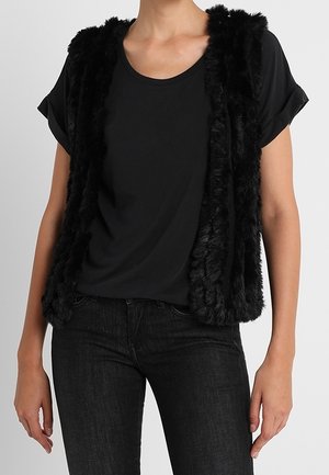 Frau trägt ein schwarzes Kurzarm-T-Shirt und eine ärmellose schwarze Kunstpelzweste mit enganliegenden schwarzen Jeans vor einem schlichten Hintergrund.