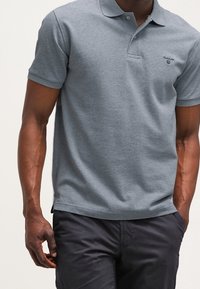 Polo gris en coton doux, avec un col, des manches courtes et un petit logo sur le côté gauche, associé à un pantalon foncé.
