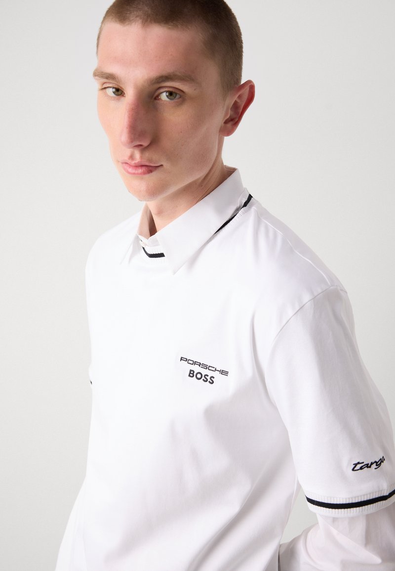 Camicia bianca con colletto e polsini neri a contrasto, loghi ricamati sul petto e sulla manica, e una trama di cotone morbido.