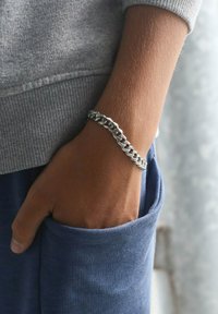Zilveren schakelarmband om een pols, met in elkaar grijpende schakels. De pols is gedeeltelijk zichtbaar in een grijze sweatshirt en een blauwe zak.