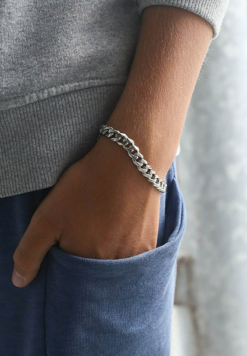 Zilveren schakelarmband om een pols, met in elkaar grijpende schakels. De pols is gedeeltelijk zichtbaar in een grijze sweatshirt en een blauwe zak.