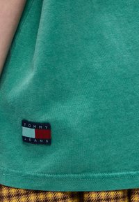 Κοντινό πλάνο υφάσματος σε τιρκουάζ χρώμα με το λογότυπο Tommy Jeans πάνω από ένδυμα με κίτρινο και καφέ καρό μοτίβο.