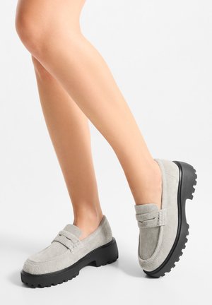 Mocassins en daim gris avec une semelle épaisse en caoutchouc noir. Le design présente des détails à double surpiqûre et un bout plat et arrondi.