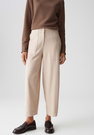 Mujer vistiendo pantalones beige de pierna ancha, una blusa marrón texturizada de manga larga y mocasines marrón oscuro, de pie contra un fondo liso.
