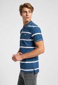 Polo a righe blu con strisce bianche, colletto, due bottoni e spacchi laterali. Il tessuto appare morbido, design aderente. Piccolo logo sul lato sinistro del petto.