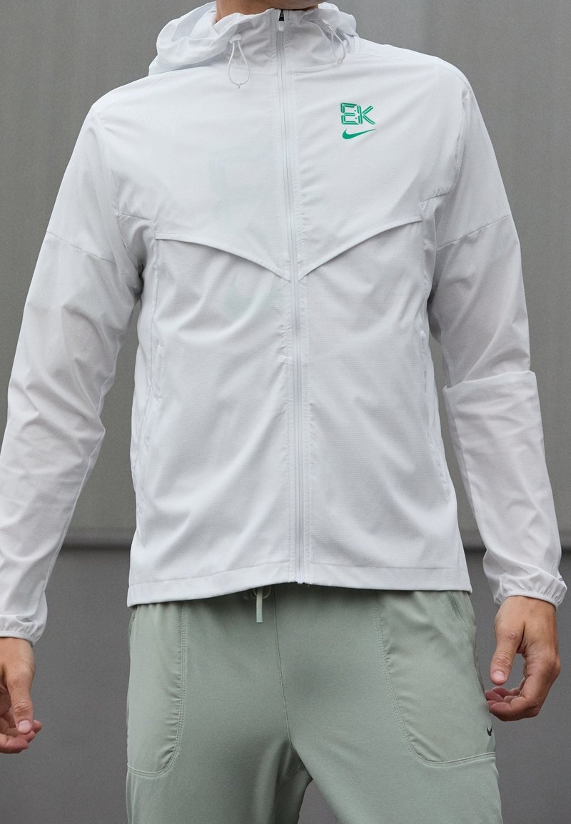 Veste blanche légère avec capuche, présentant un design de poitrine divisé et des accents du logo Nike verts, associée à un pantalon vert clair.