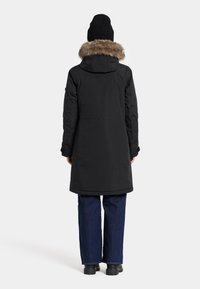 Sort parka-jakke med faux fur-kant på hætten, lang længde og en glat tekstur. Sammen med mørke denim jeans og sorte støvler.
